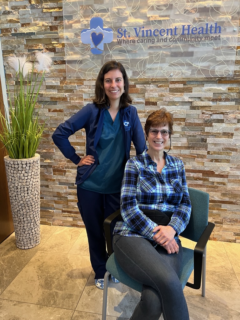 Wound Care nurse, Lindsy Coon (standing) is pictured with her patient Marianne Maes (sitting) in front of the St. Vincent Health Logo wall.